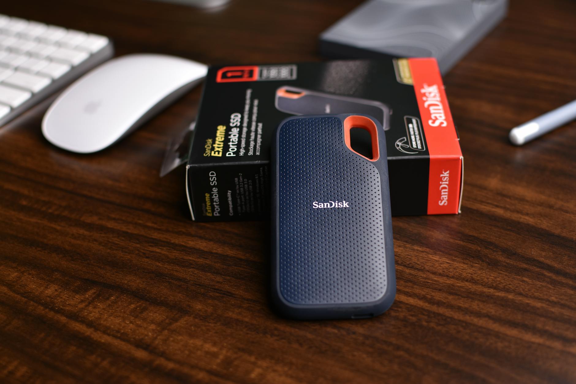 Przenośny dysk SSD SanDisk obok pudełka i myszy na drewnianym biurku